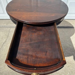 May include: A dark brown, round side table with a drawer. The table has a polished finish, and the drawer is open, revealing the interior. The drawer has a brass-colored handle. The table is approximately 24 inches in diameter.