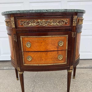 May include: An antique, oval-shaped chest of drawers with a green marble top. The chest features two drawers with brass hardware and decorative gold accents. The wood has a warm, reddish-brown finish with lighter wood panels. The chest stands on four slender, tapered legs.
