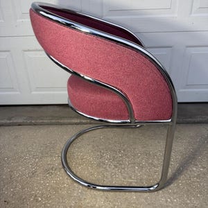 May include: A modern chair with a curved, chrome-finished metal frame. The seat and backrest are upholstered in a textured, dusty rose fabric. The chair has a unique, open design.