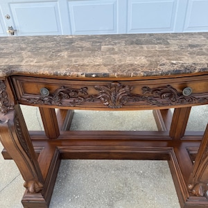May include: A brown marble-topped console table with ornate carved details. The table features a curved front with a drawer and decorative hardware. The legs are intricately carved and supported by a lower stretcher, creating a classic, elegant look.