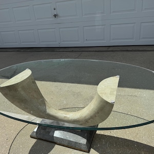 May include: An oval glass coffee table featuring a sculptural, beige stone base with a curved, U-shaped design. The clear glass top is supported by the stone structure, which rests on a rectangular, dark gray stone pedestal base at the bottom.