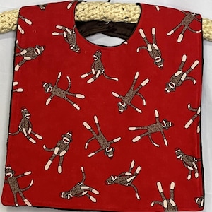 May include: Red baby bib with a repeating pattern of brown sock monkey figures with white accents. The bib has a rounded neckline and a black backing. The bib is hanging on a beige rope hanger.