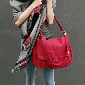 May include: A red canvas shoulder bag with a flap closure and adjustable strap. The bag is paired with a gray scarf with a red and black plaid pattern. The person is wearing a gray top and blue jeans.