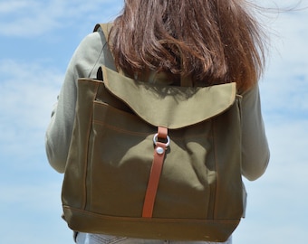 May include: An olive green canvas backpack with a flap closure and brown leather straps. The backpack is worn by a person with long brown hair, standing against a blue sky with white clouds. The bag has a metal buckle.
