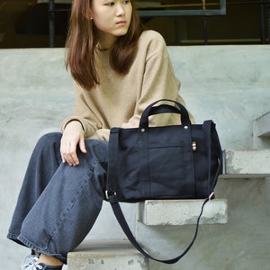 Puede incluir: Un bolso tote de lona negra con una correa para el hombro y asas superiores. El bolso tiene un bolsillo frontal y un cierre de cremallera. El texto "Place Where by APW" es visible en el fondo.