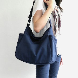 May include: A navy blue shoulder bag with a black strap is carried over the shoulder. The bag has a rectangular shape with two front pockets. The person is wearing blue jeans and tan leather boots.