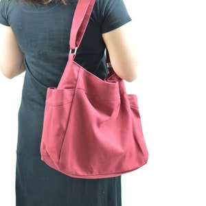 May include: A burgundy tote bag with a shoulder strap. The bag has two exterior pockets and a large main compartment. The strap is attached to the bag with silver metal rings. The bag is made of a sturdy fabric.