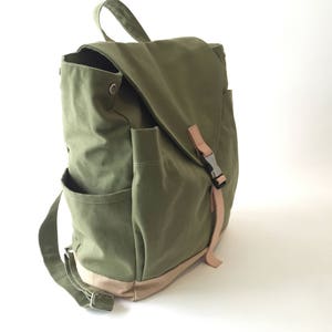 May include: Olive green canvas backpack with a flap closure and side pockets. Features a tan base and straps, with a metal buckle closure. The bag has a top handle and adjustable shoulder straps for comfortable carrying.