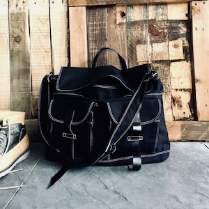 May include: Black messenger bag with white stitching and multiple pockets. The bag has a top handle and a shoulder strap. The background is a wooden wall and a stone floor. A pair of boots is visible on the left.