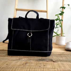 May include: A black canvas backpack with a flap closure and a top handle. The bag features white stitching and a leather strap with a silver ring closure. The bag is sitting on a wooden surface.