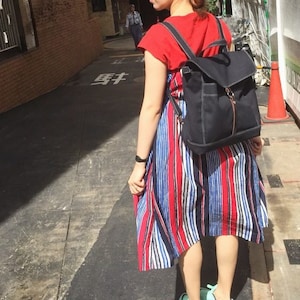 May include: A black backpack with a flap closure and a gold zipper is worn over a red top and a striped dress with red, white, and blue vertical stripes. The person is wearing blue and orange sneakers.