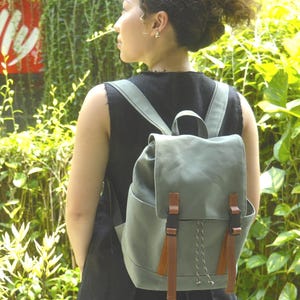 May include: A sage green backpack with brown leather accents and straps. The backpack has a flap closure with two brown straps and black buckles. The bag has a drawstring closure and is worn on the back.