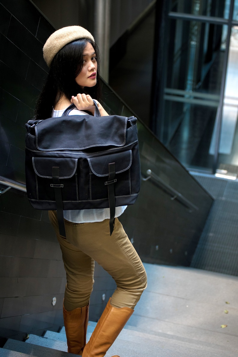 May include: A black messenger bag with white stitching and two front pockets. The bag has a flap closure with two black straps. The person in the image is wearing a beige beret, a white shirt, khaki pants, and brown boots.