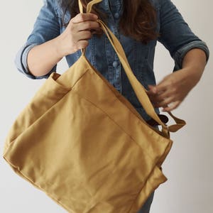 May include: A large, mustard yellow canvas tote bag with two long straps. The bag has a large pocket on the front and is made of a soft, durable fabric.