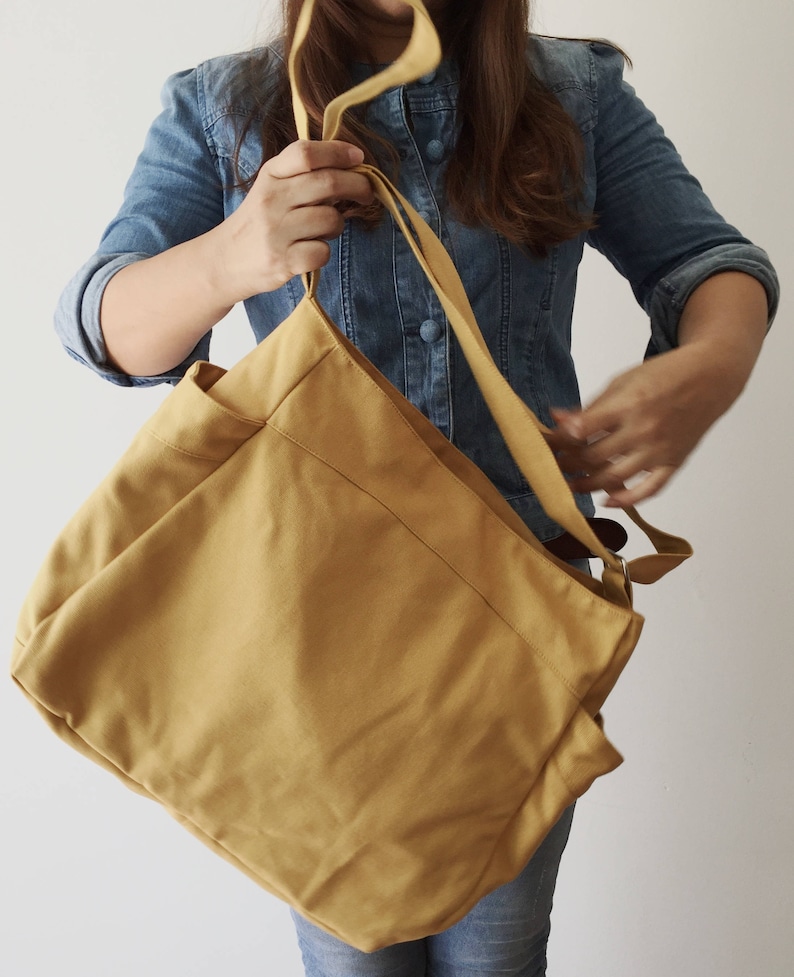 May include: A large, mustard yellow canvas tote bag with two long straps. The bag has a large pocket on the front and is made of a soft, durable fabric.