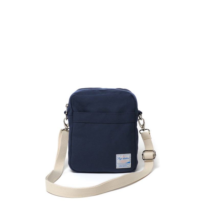 May include: A navy blue canvas crossbody bag with a cream-colored adjustable strap. The bag has a rectangular shape with a small rectangular label on the front. The label has text and a graphic. The bag is designed to be worn over the shoulder.