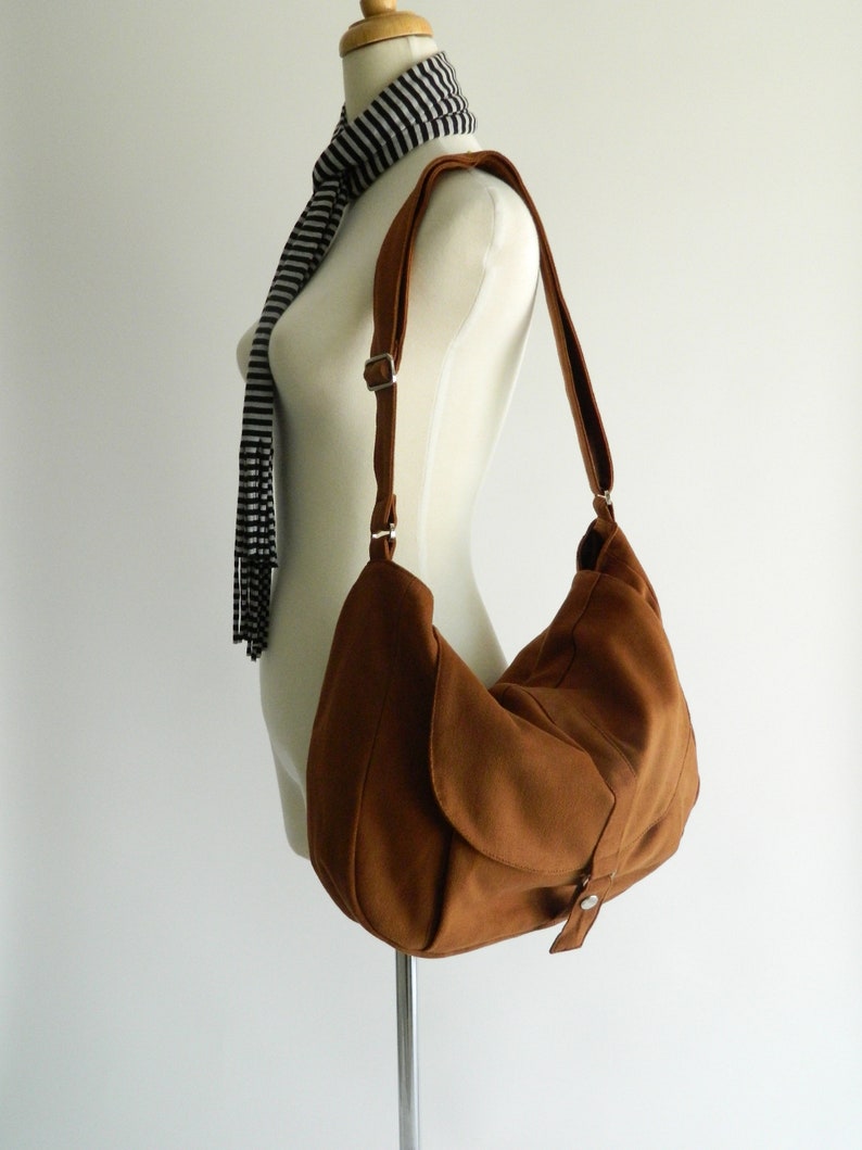 May include: A brown shoulder bag with a curved design and adjustable strap. The bag is displayed on a mannequin, showcasing its shape and style. The bag has a flap closure with a metal button.