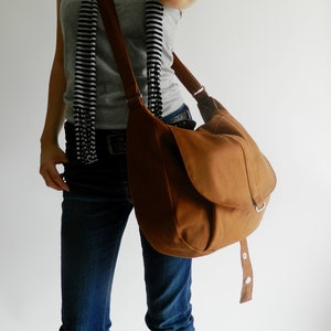 May include: A brown shoulder bag with a flap closure and adjustable strap. The bag is made of a soft, suede-like material. The bag is worn over the shoulder. The person is wearing blue jeans, a gray tank top, and a black and white striped scarf.