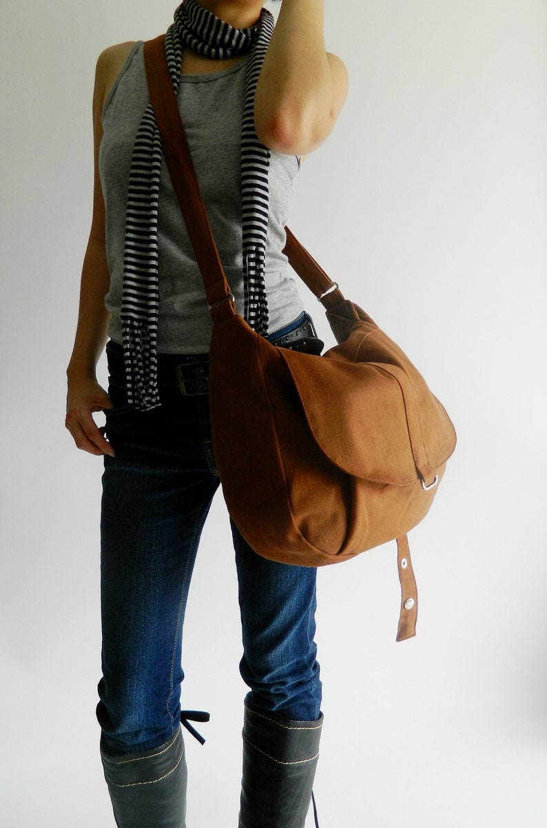 May include: A brown shoulder bag with a flap closure and adjustable strap. The bag is made of a soft, suede-like material. The bag is worn over the shoulder. The person is wearing blue jeans, a gray tank top, and a black and white striped scarf.