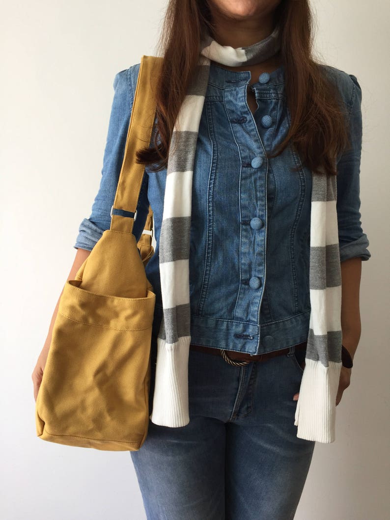 May include: A mustard yellow canvas tote bag with a long strap. The bag is being held by a person wearing a denim shirt and jeans.