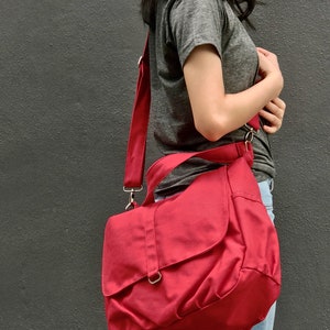May include: A red messenger bag with a flap closure and adjustable shoulder strap. The bag is made of a durable fabric and features silver-colored metal hardware. The bag is designed to be worn over the shoulder.