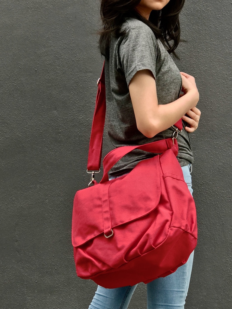 May include: A red messenger bag with a flap closure and adjustable shoulder strap. The bag is made of a durable fabric and features silver-colored metal hardware. The bag is designed to be worn over the shoulder.