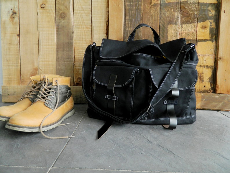May include: A black canvas messenger bag with white stitching and multiple pockets, alongside a pair of tan and gray leather boots with laces. The bag has a shoulder strap and leather accents. The background is a wooden wall.