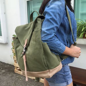 May include: An olive green canvas backpack with a flap closure and a light brown base. The backpack has a top handle and adjustable straps. A pink strap secures the flap. The bag is carried over the shoulder.