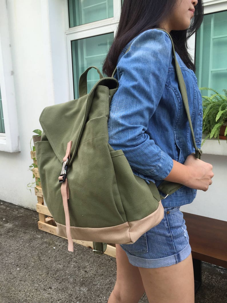May include: An olive green canvas backpack with a flap closure and a light brown base. The backpack has a top handle and adjustable straps. A pink strap secures the flap. The bag is carried over the shoulder.