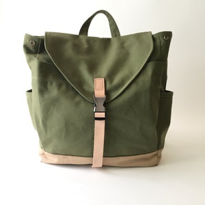 May include: Olive green canvas backpack with a tan bottom and flap closure. Features a tan strap with a metal buckle and a top handle. Includes two side pockets and a front pocket.