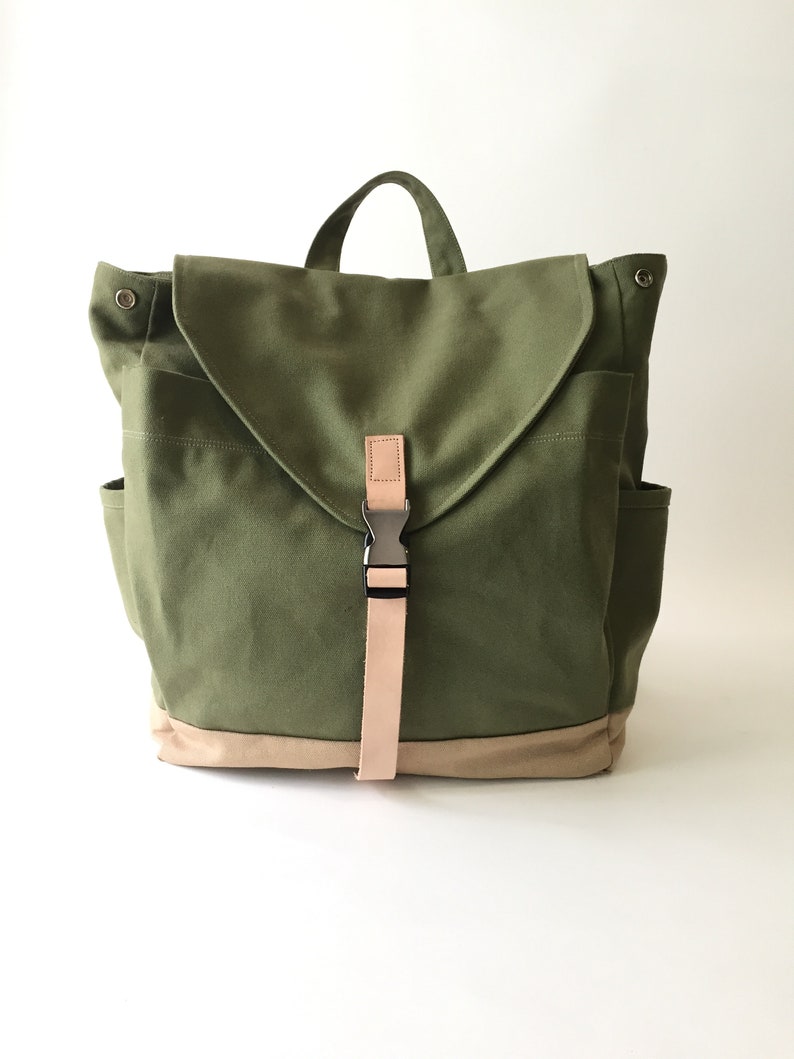 May include: Olive green canvas backpack with a tan bottom and flap closure. Features a tan strap with a metal buckle and a top handle. Includes two side pockets and a front pocket.
