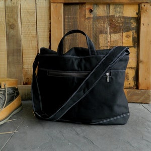 May include: Black canvas tote bag with white stitching and a shoulder strap. The bag has a zipper closure and two handles. The bag is sitting on a gray surface in front of a wooden background.