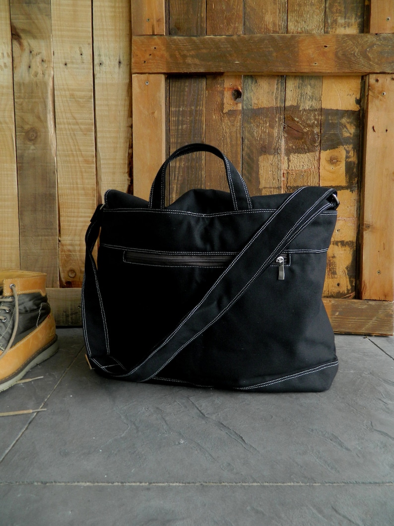 May include: Black canvas tote bag with white stitching and a shoulder strap. The bag has a zipper closure and two handles. The bag is sitting on a gray surface in front of a wooden background.