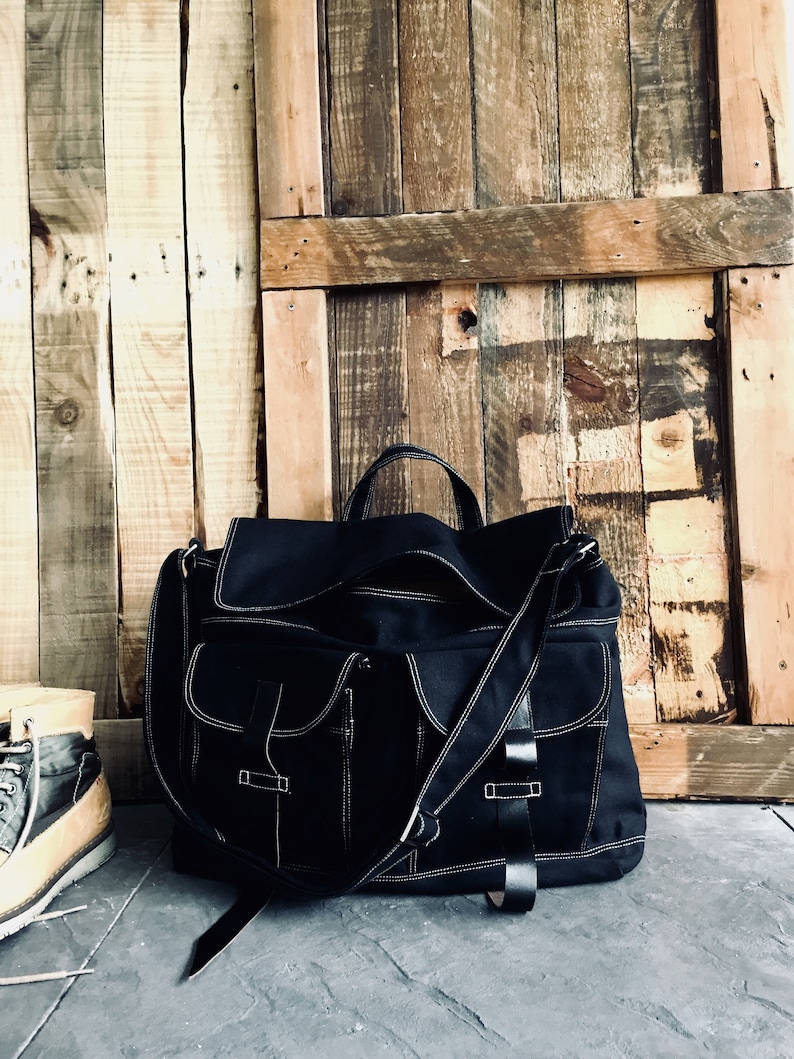 May include: Black messenger bag with white stitching and multiple pockets. The bag has a top handle and a shoulder strap. The background is a wooden wall and a stone floor. A pair of boots is visible on the left.