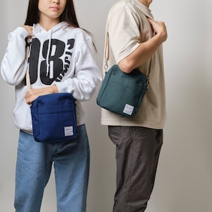 May include: Two people are shown wearing crossbody bags. One bag is navy blue, and the other is teal. Both bags have a rectangular shape with a front pocket and a small label. The bags are worn with casual outfits.