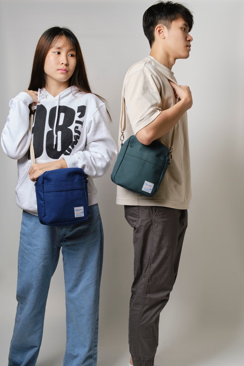 May include: Two people are shown wearing crossbody bags. One bag is navy blue, and the other is teal. Both bags have a rectangular shape with a front pocket and a small label. The bags are worn with casual outfits.
