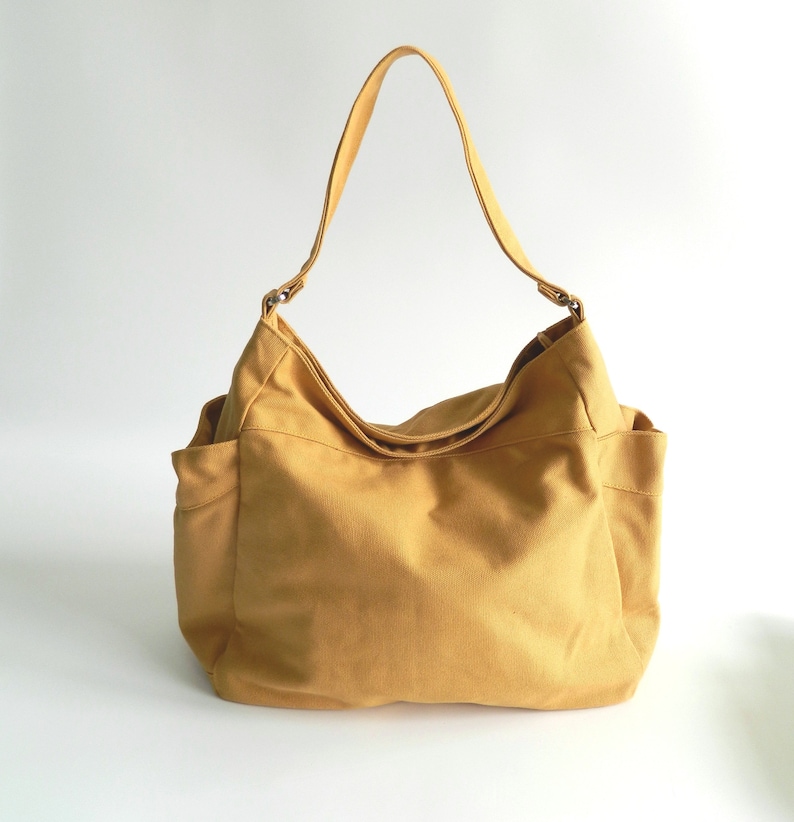 May include: A large, mustard yellow canvas tote bag with a single strap. The bag has a slouchy, casual look and is made of a durable, natural fabric.