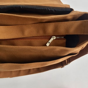K&ouml;nnte beinhalten: Nahaufnahme einer braunen Canvas-Umh&auml;ngetasche. Die Tasche verf&uuml;gt &uuml;ber eine Rei&szlig;verschlusstasche mit silbernem Rei&szlig;verschluss, einen verstellbaren braunen Riemen und ein schwarzes Innenfutter. Die Tasche ist zum Tragen von Gegenst&auml;nden konzipiert.