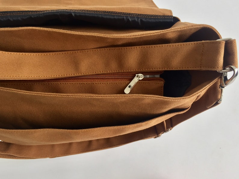 K&ouml;nnte beinhalten: Nahaufnahme einer braunen Canvas-Umh&auml;ngetasche. Die Tasche verf&uuml;gt &uuml;ber eine Rei&szlig;verschlusstasche mit silbernem Rei&szlig;verschluss, einen verstellbaren braunen Riemen und ein schwarzes Innenfutter. Die Tasche ist zum Tragen von Gegenst&auml;nden konzipiert.
