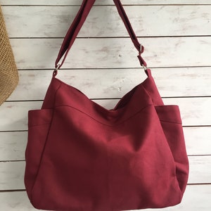 May include: A burgundy tote bag with a shoulder strap. The bag has two exterior pockets and is hanging against a white wooden background. The bag is made of a solid, textured fabric.