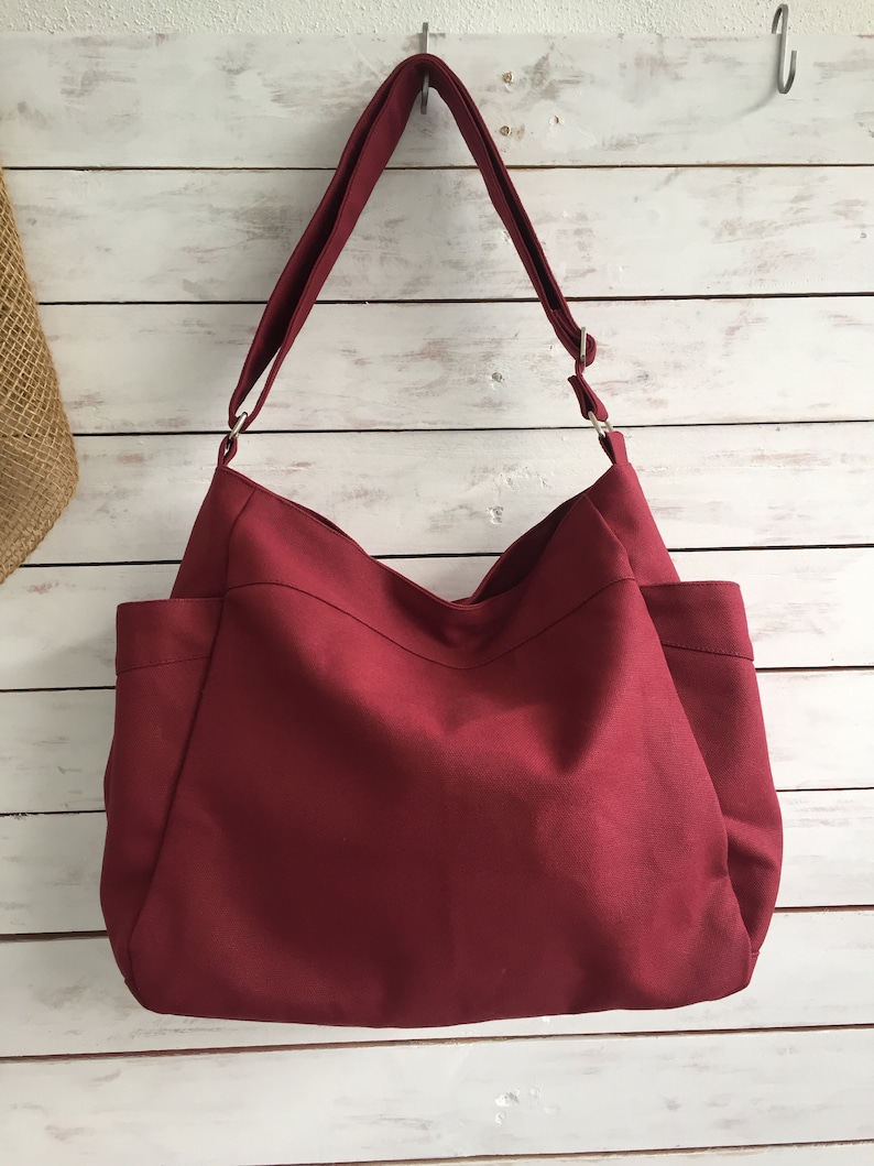 May include: A burgundy tote bag with a shoulder strap. The bag has two exterior pockets and is hanging against a white wooden background. The bag is made of a solid, textured fabric.