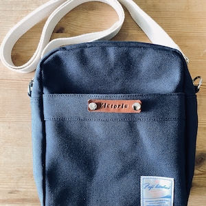 May include: A navy blue canvas crossbody bag with a cream-colored adjustable strap. The bag features a front pocket and a small leather tag with the name "Victoria". A label with Japanese text is on the lower right corner.