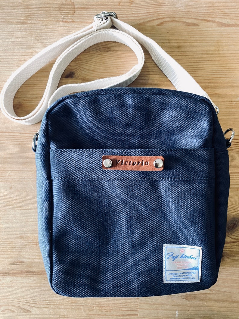 May include: A navy blue canvas crossbody bag with a cream-colored adjustable strap. The bag features a front pocket and a small leather tag with the name "Victoria". A label with Japanese text is on the lower right corner.