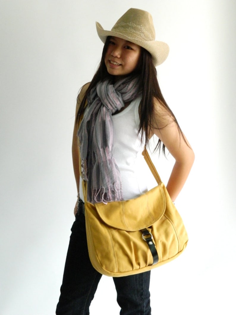 May include: A mustard yellow messenger bag with a black buckle closure and a long shoulder strap. The bag is made of a soft material and has a flap closure. The bag is worn with a white tank top, a scarf, and a straw cowboy hat.