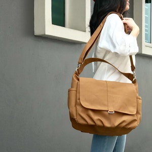 K&ouml;nnte beinhalten: Eine hellbraune Messenger-Tasche mit Klappe und verstellbarem Schultergurt. Die Tasche hat zwei Seitentaschen und eine silberfarbene Schnalle. Die Tasche ist aus robustem Material gefertigt und wird &uuml;ber der Schulter getragen.