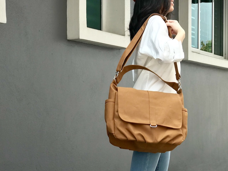 K&ouml;nnte beinhalten: Eine hellbraune Messenger-Tasche mit Klappe und verstellbarem Schultergurt. Die Tasche hat zwei Seitentaschen und eine silberfarbene Schnalle. Die Tasche ist aus robustem Material gefertigt und wird &uuml;ber der Schulter getragen.