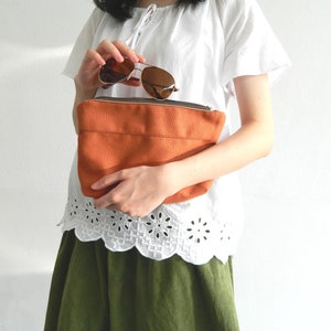 Op de afbeelding: Een vrouw houdt een bruine canvas tas vast met een zonnebril erin. De tas heeft een witte rits en is gemaakt van een zachte, textuurrijke stof. De vrouw draagt een wit blouse met een kantversiering en een groene rok.