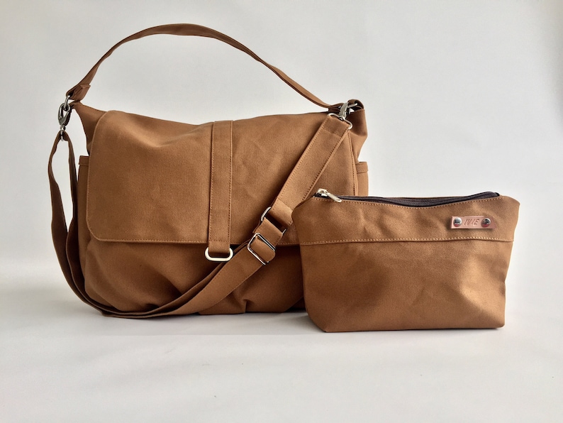 K&ouml;nnte beinhalten: Eine braune Messenger Bag und eine passende kleine Tasche. Die Messenger Bag hat einen Klappverschluss, einen verstellbaren Schultergurt und silberfarbene Beschl&auml;ge. Die Tasche hat einen Rei&szlig;verschluss und ein kleines Metallschild. Beide sind aus robustem Canvas-Material.
