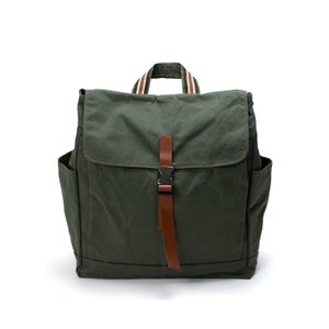 Puede incluir: Una mochila verde oliva con cierre de solapa y correa de cuero marrón. La mochila tiene bolsillos laterales y un asa a rayas. La bolsa está hecha de un material de lona duradero.