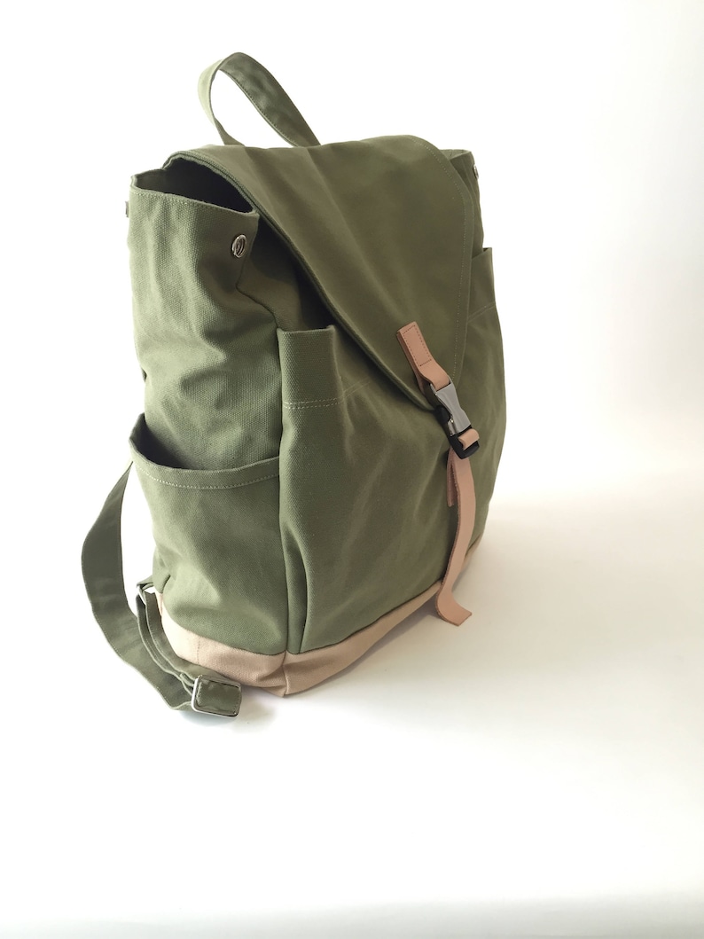 May include: Olive green canvas backpack with a flap closure and side pockets. Features a tan base and straps, with a metal buckle closure. The bag has a top handle and adjustable shoulder straps for comfortable carrying.